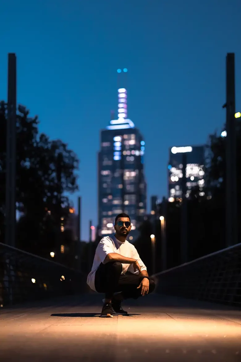 SPE3D, the talented artist, kneeling on a bridge against the backdrop of a city skyline. Soft lighting accentuates the portrait, creating a captivating ambiance in dark azure and white tones.