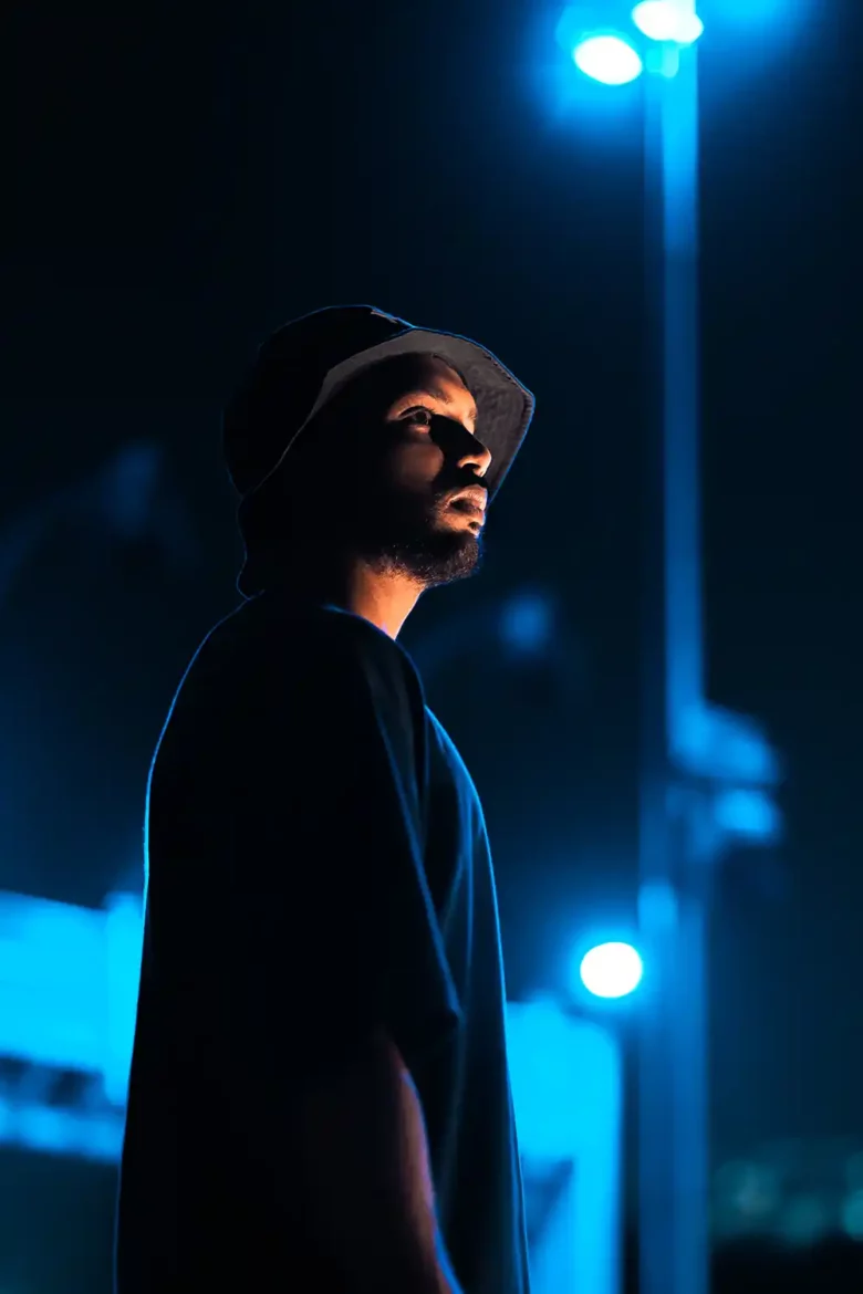 Ethereal image of the artist wearing a black cap amidst vibrant stage backdrops, with stunning light effects and shallow depth of field. Moody monotones add a captivating atmosphere.