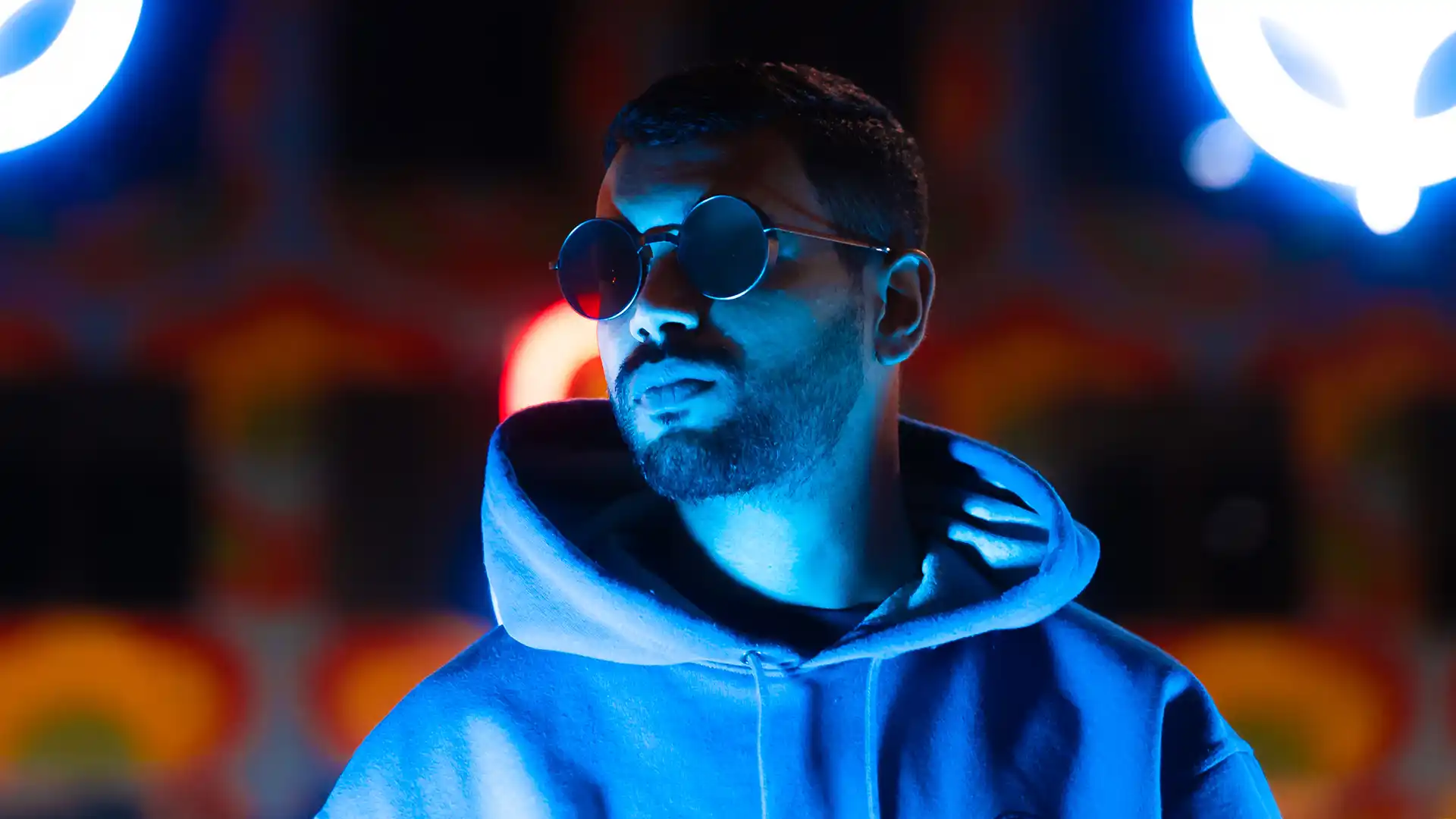 Artist SPE3D standing confidently in front of vibrant neon lights, wearing sunglasses.