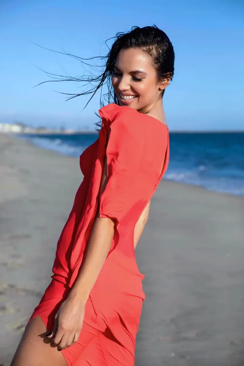 Angel Carra exudes joy and elegance in a flowing red dress, smiling amidst the beach's gentle breeze during a captivating photoshoot.