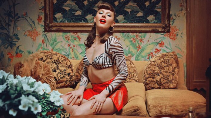 Meresha with a bold red lipstick and green eyeshadow sits on a mustard-yellow couch, wearing a zebra print top and red shorts, exuding confidence.