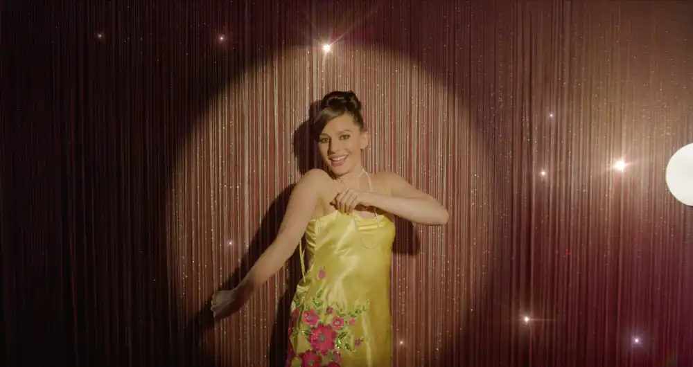 Smiling Meresha in a floral yellow dress dancing in front of a shimmering curtain with heart-shaped light flare.