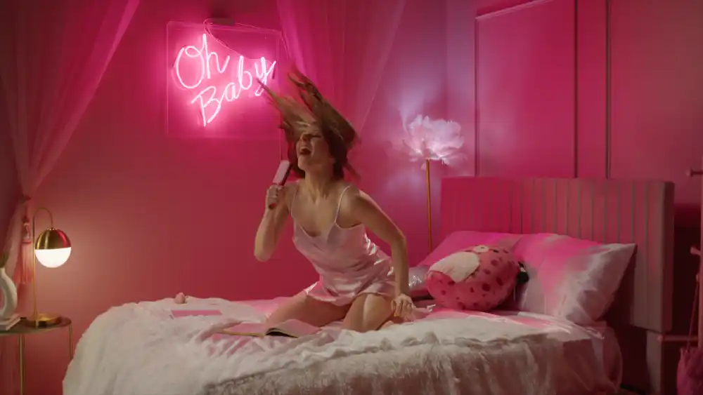Energetic Meresha singing into a hairbrush, with her hair flying, in a pink-themed room with a neon sign reading "Oh Baby."