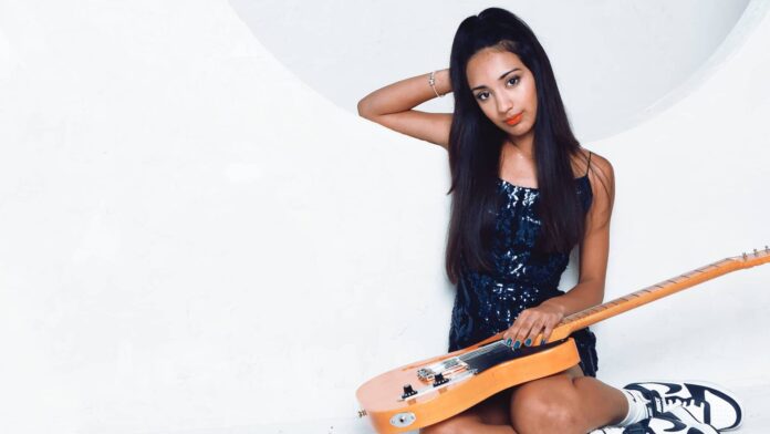 Young female artist Sophia Sheth with a guitar sitting against a white backdrop