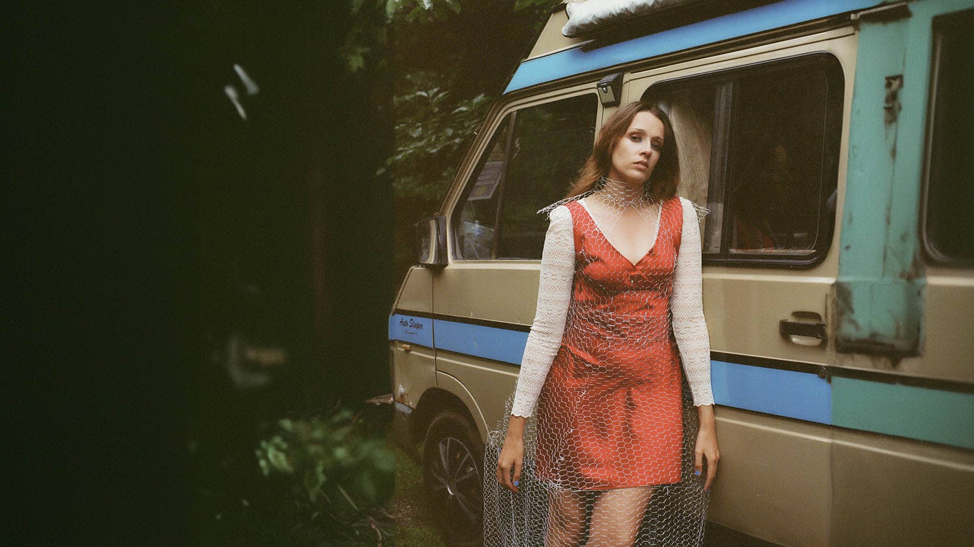 Singer-songwriter Sophie Kilburn stands beside a vintage van, clad in a red dress layered with a white lace top and a netted overlay.