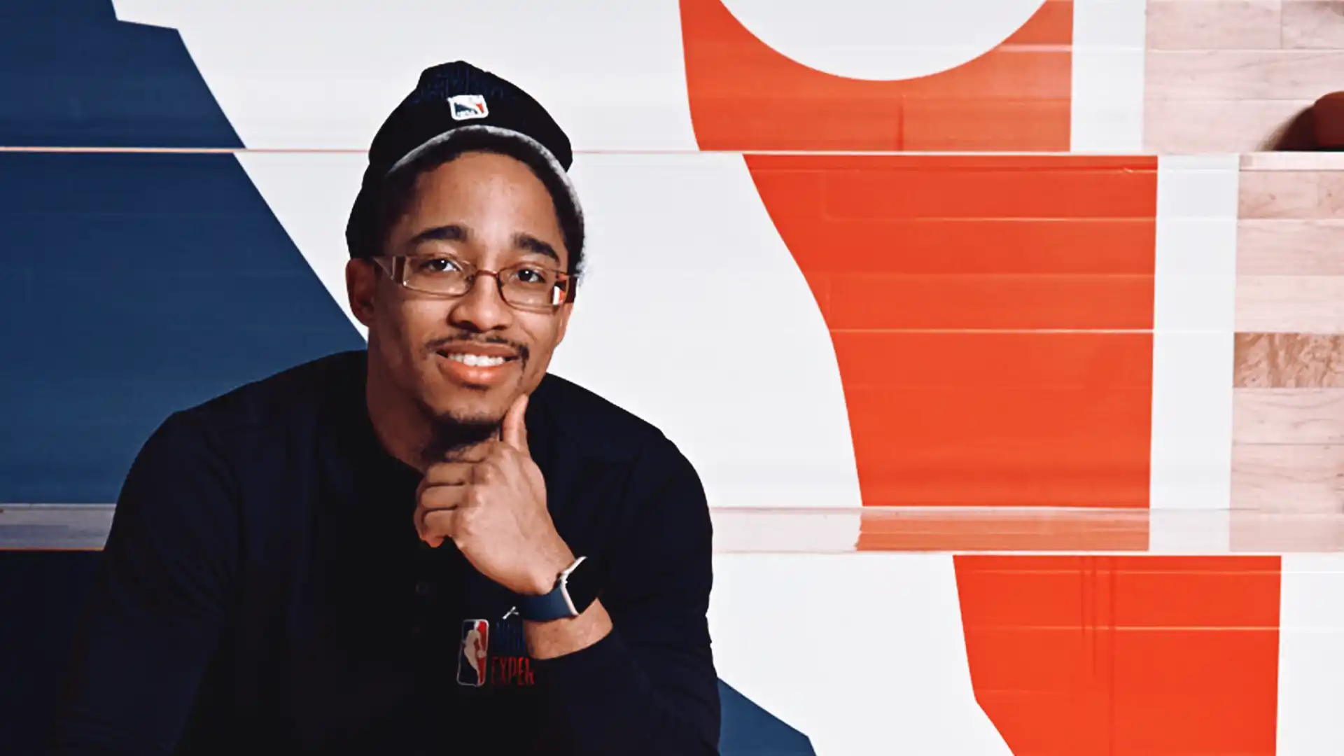 Smiling man in a black shirt, seated in front of an NBA logo backdrop.