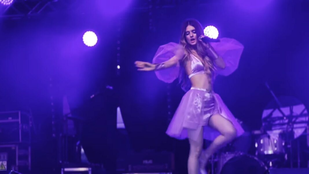 Juli Chan performs on stage during The Posh Tour, dressed in a lilac outfit, with bright vertical lights in the background.